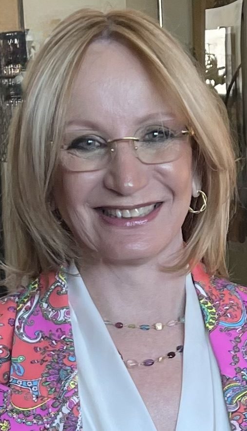 Smiling woman with glasses and blonde hair wearing a colorful outfit.