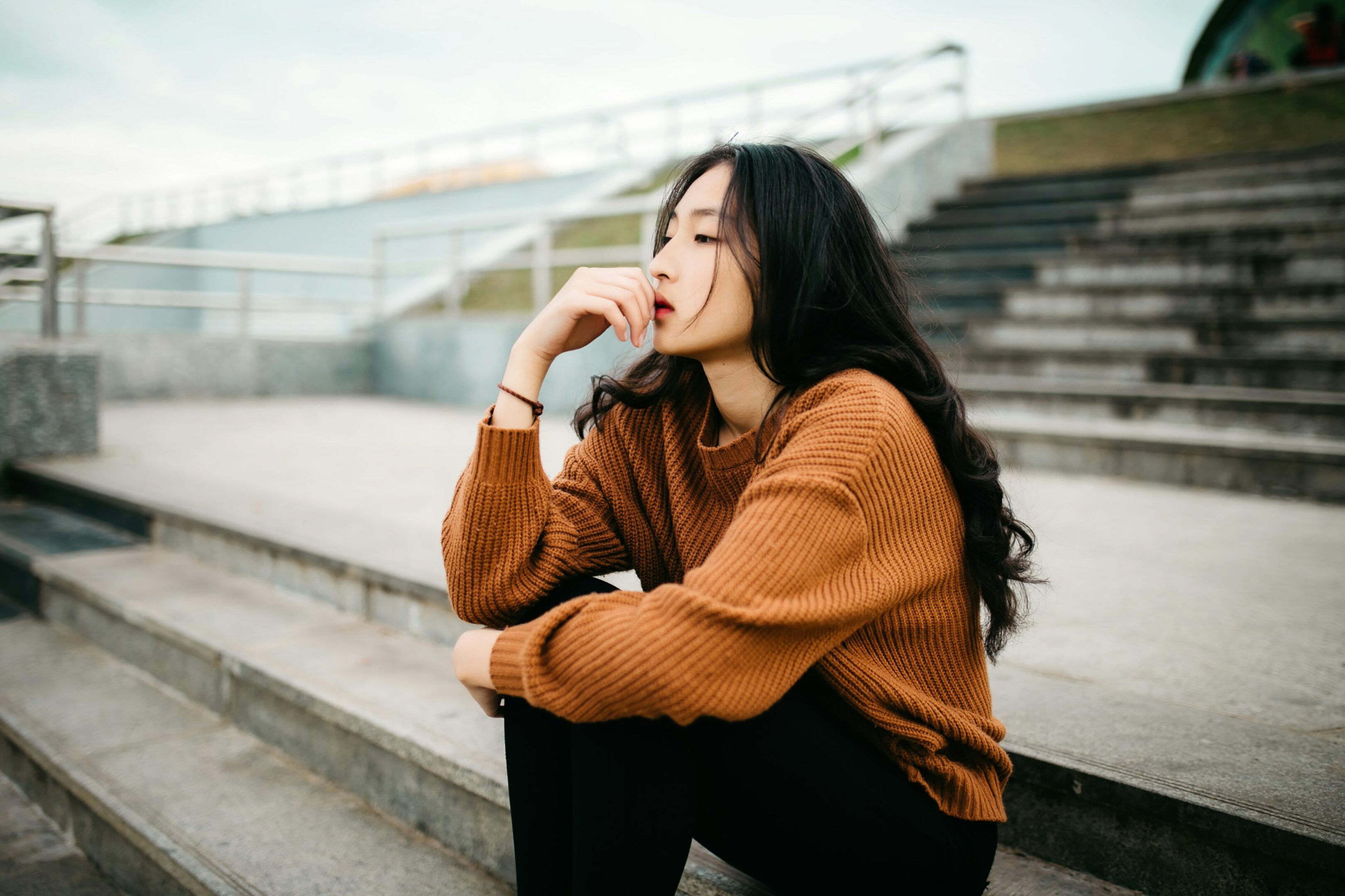 Woman in a brown sweater sitting thoughtfully on outdoor stairs.