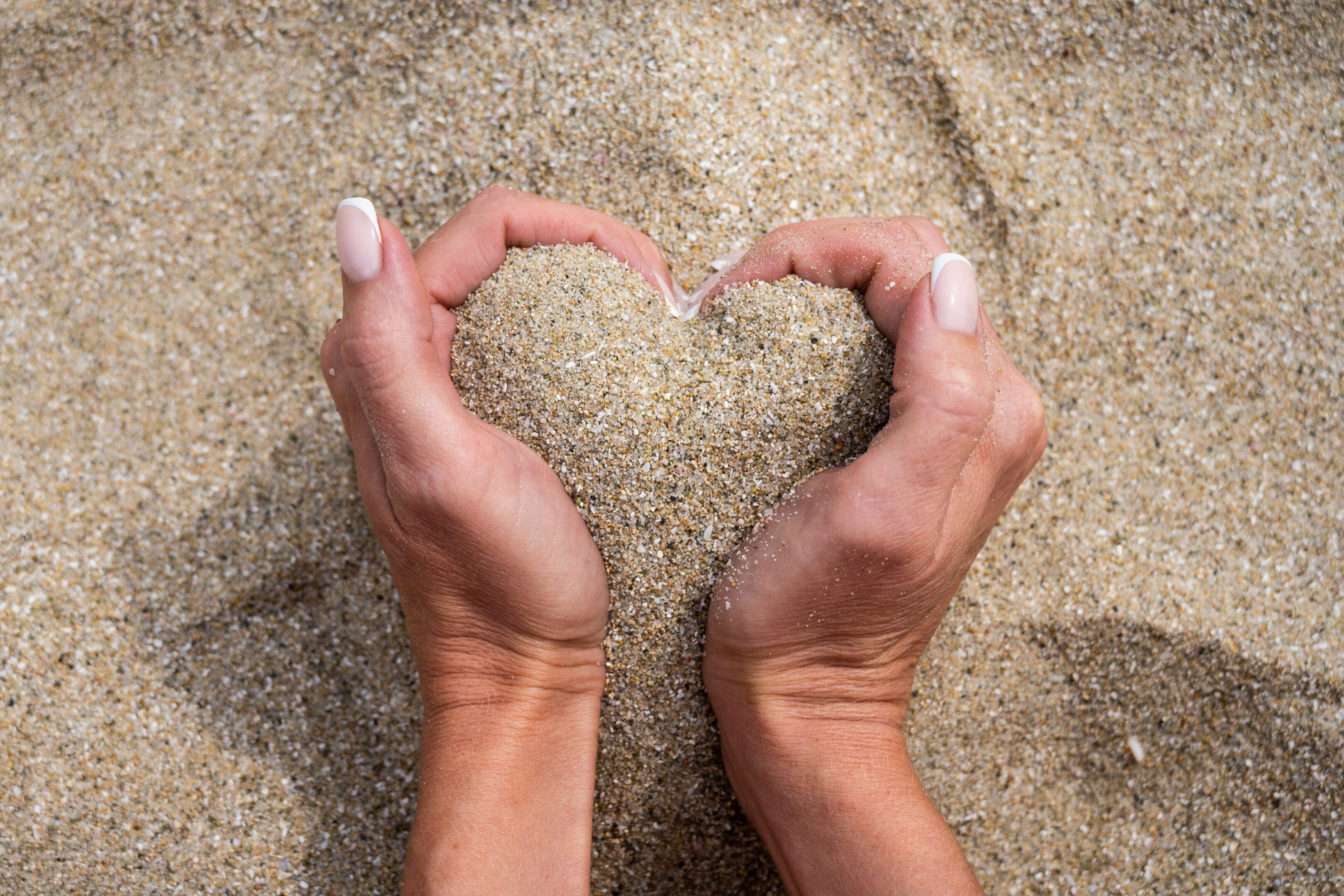 Hands forming a heart shape with sand inside on the beach.